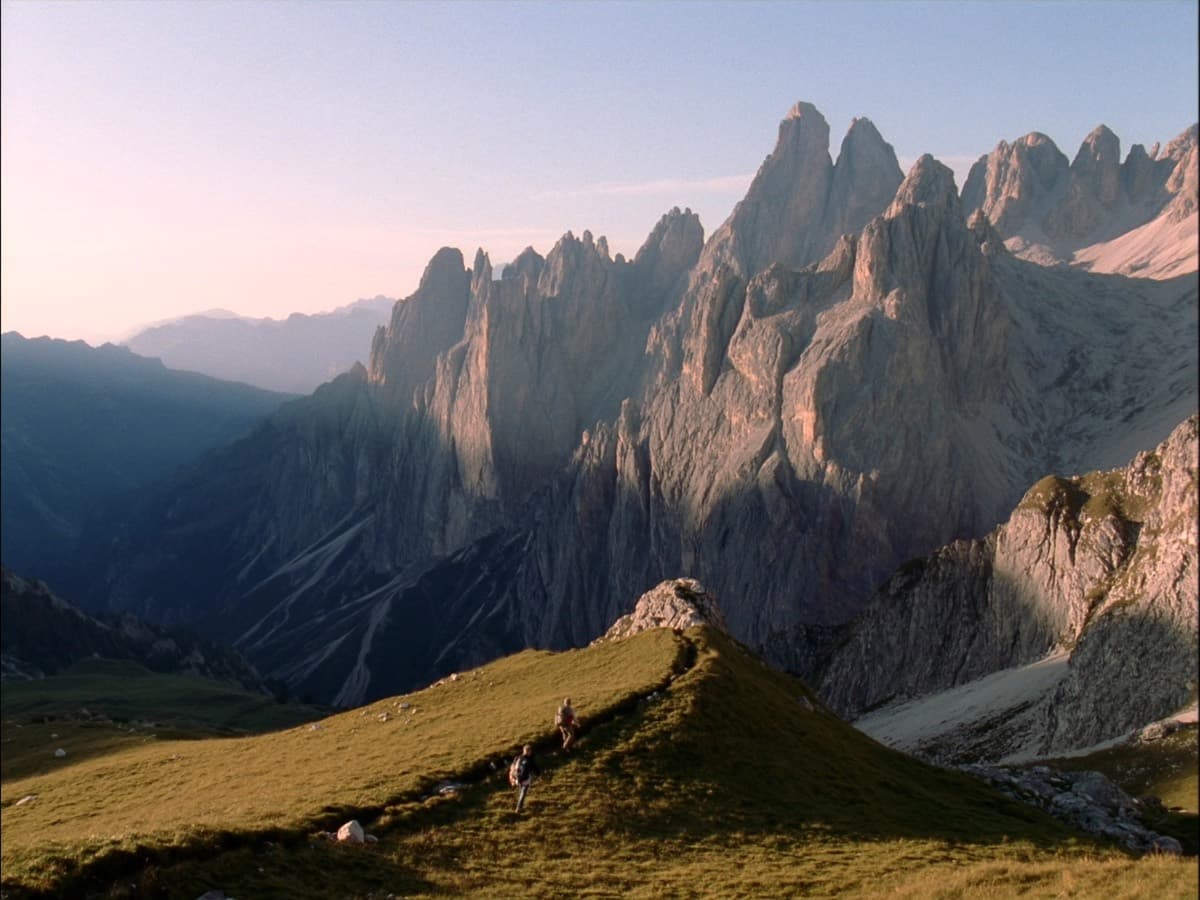 Two hikers on a high alpine meadow trail below the pale limestone peaks of the Dolomites